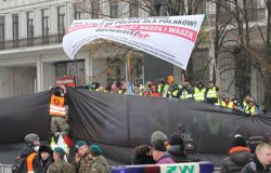 Mężczyzna w kamizelce z logiem „Straż Marszu Niepodległości” mocuje czarne płachty, które mają zasłonić pikietę „Obywatele RP”.